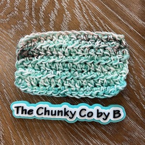 May include: A rectangular, hand-crocheted item in shades of teal, white, and brown. The item is labeled with the text "The Chunky Co by B" in a teal and white design.