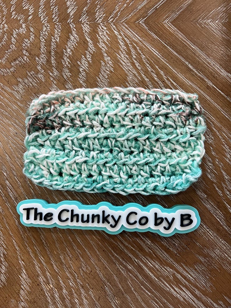 May include: A rectangular, hand-crocheted item in shades of teal, white, and brown. The item is labeled with the text "The Chunky Co by B" in a teal and white design.
