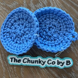 May include: Two round, sky blue, crocheted items with a loop on one side. The items are made of thick yarn and are on a wooden surface. A label with the text "The Chunky Co by B" is below the items.
