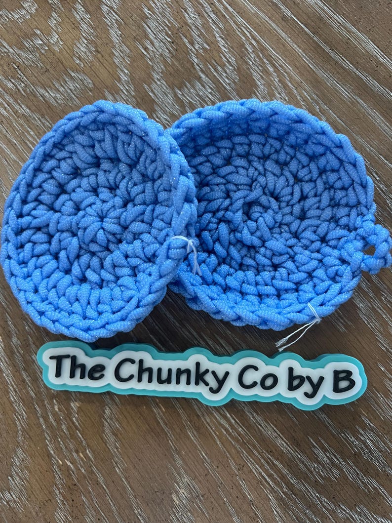 May include: Two round, sky blue, crocheted items with a loop on one side. The items are made of thick yarn and are on a wooden surface. A label with the text "The Chunky Co by B" is below the items.