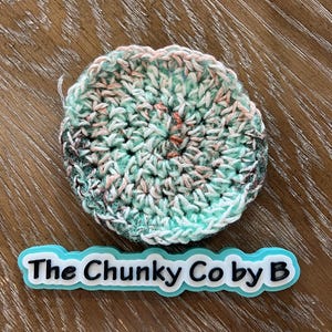 May include: A circular, crocheted item in teal, white, pink, and brown hues. It rests on a wooden surface. The text "The Chunky Co by B" is displayed on a blue and white label.