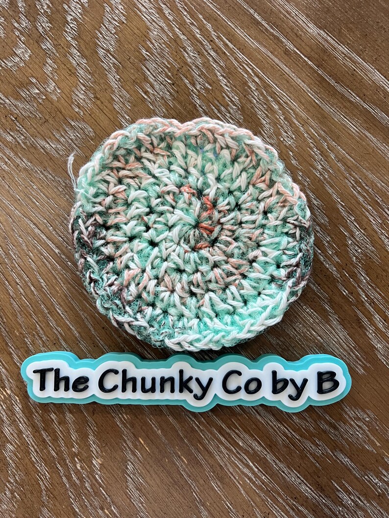 May include: A circular, crocheted item in teal, white, pink, and brown hues. It rests on a wooden surface. The text "The Chunky Co by B" is displayed on a blue and white label.