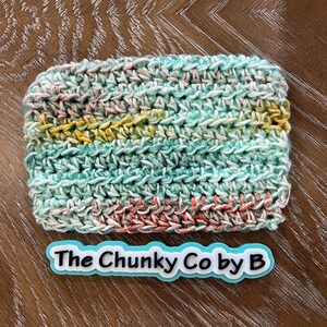 May include: A rectangular, hand-crocheted item in shades of teal, pink, yellow, and white. The text "The Chunky Co by B" is displayed on a light blue and white label below the item. The item is on a wooden surface.