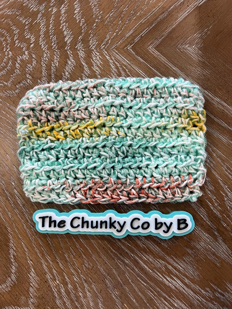 May include: A rectangular, hand-crocheted item in shades of teal, pink, yellow, and white. The text "The Chunky Co by B" is displayed on a light blue and white label below the item. The item is on a wooden surface.