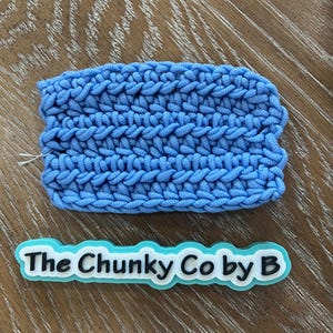May include: A light blue, chunky-knit item with a textured surface. The item is rectangular and appears to be handmade. Below the item is a white and light blue sticker with the text "The Chunky Co by B".