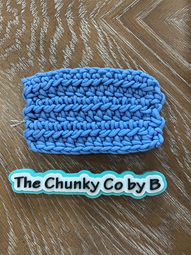 May include: A light blue, chunky-knit item with a textured surface. The item is rectangular and appears to be handmade. Below the item is a white and light blue sticker with the text "The Chunky Co by B".
