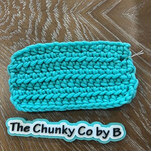 May include: A turquoise crocheted item with a thick, textured stitch pattern. The handmade item has a white thread extending from the edge. A sticker with the text "The Chunky Co by B" is below the crocheted item.