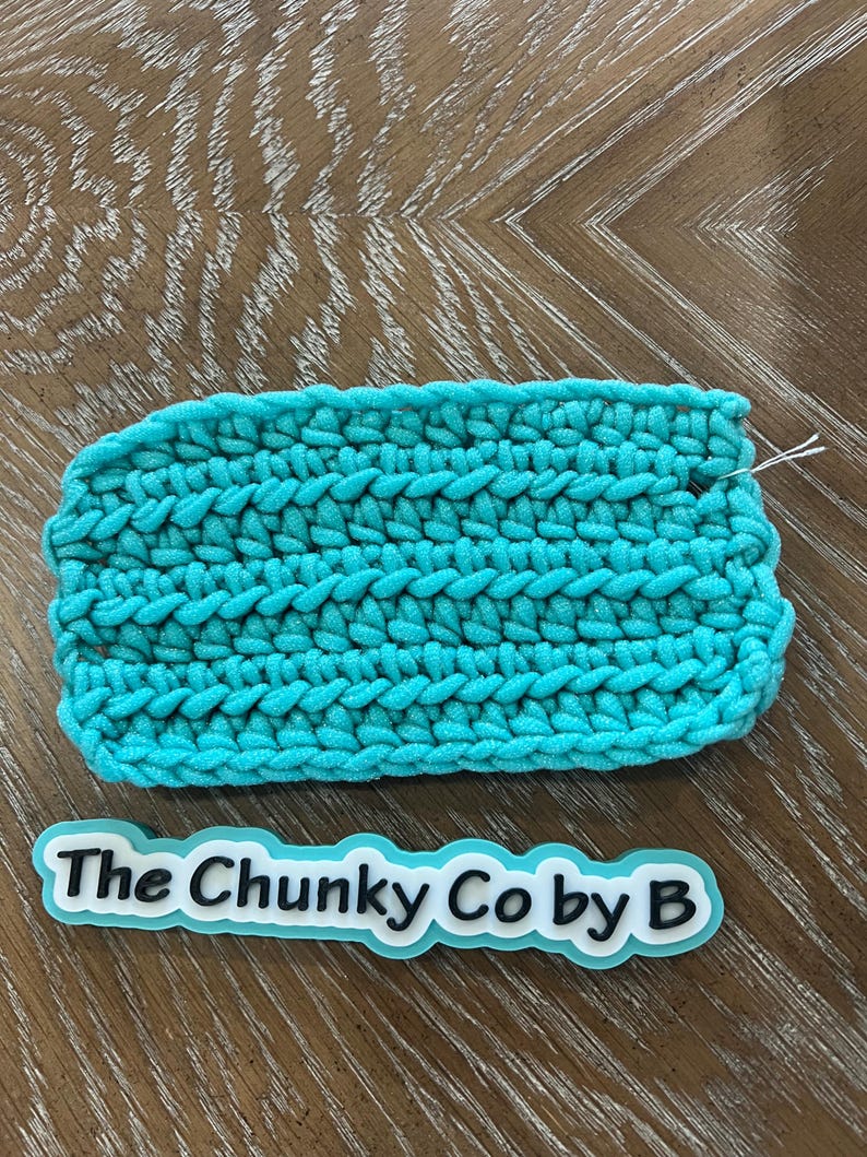 May include: A turquoise crocheted item with a thick, textured stitch pattern. The handmade item has a white thread extending from the edge. A sticker with the text "The Chunky Co by B" is below the crocheted item.