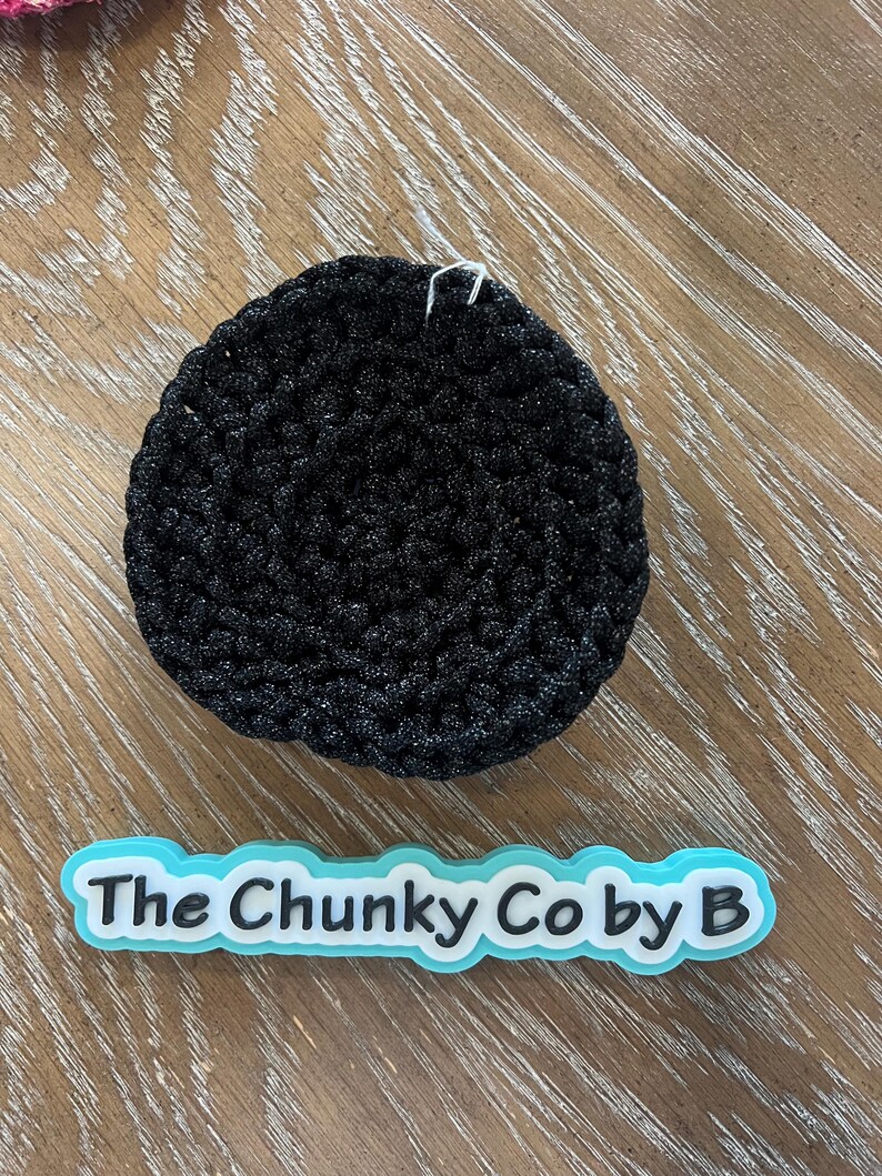 May include: A black, circular, crocheted item with a textured surface. Below it is a light blue and white logo that reads "The Chunky Co by B". The item is on a wooden surface.