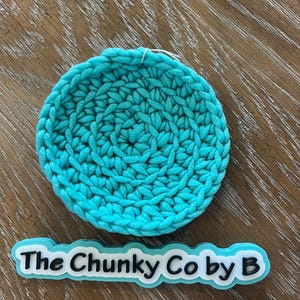 May include: A circular, turquoise crocheted coaster. The coaster is made from thick yarn, giving it a textured look. The text "The Chunky Co by B" is displayed on a white and turquoise label below the coaster.