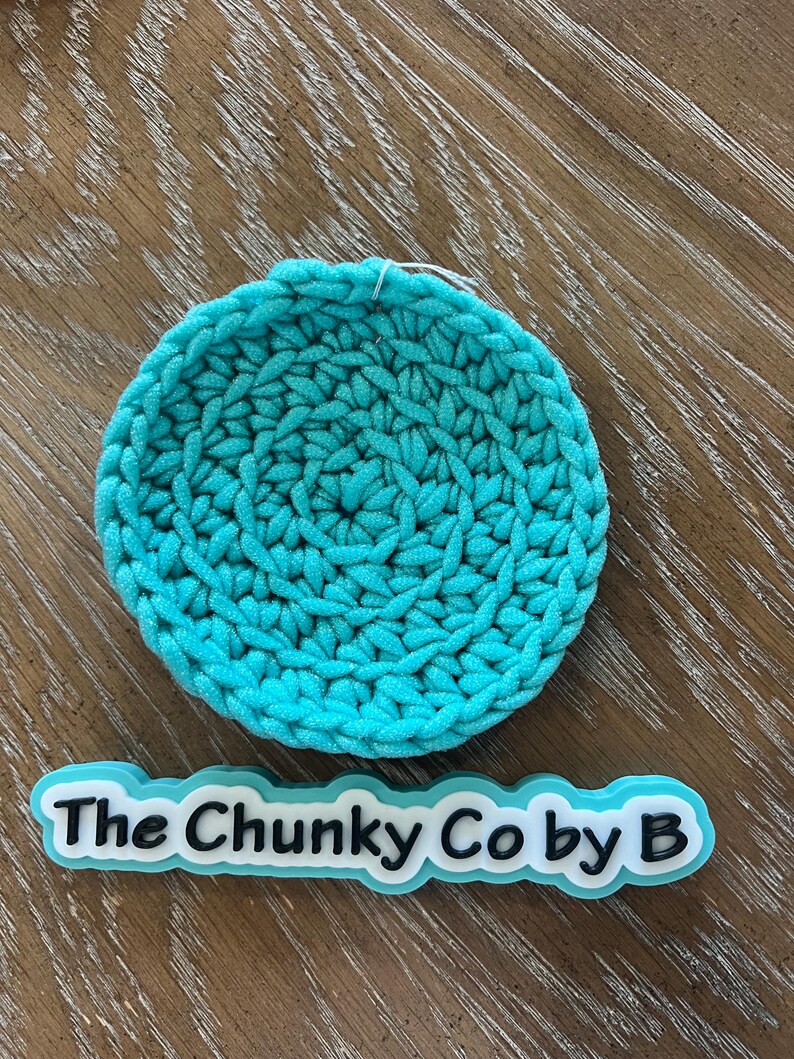 May include: A circular, turquoise crocheted coaster. The coaster is made from thick yarn, giving it a textured look. The text "The Chunky Co by B" is displayed on a white and turquoise label below the coaster.