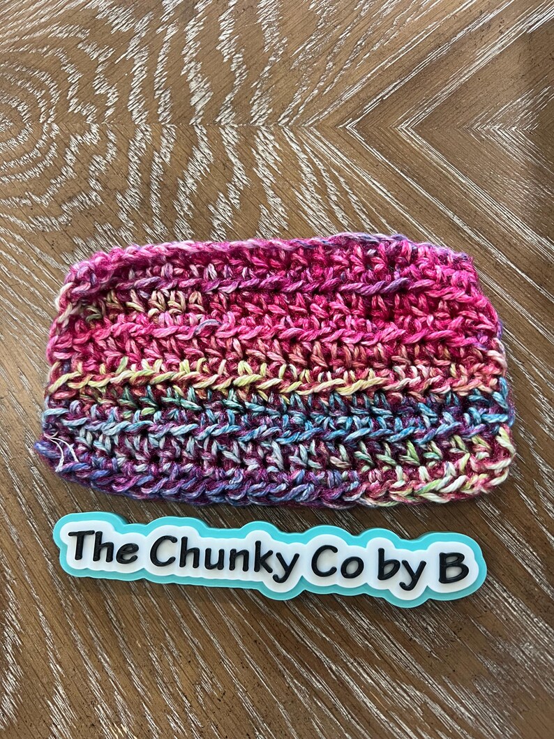 May include: A colourful, hand-crocheted beanie hat in shades of pink, purple, blue, and yellow. The hat has a textured stitch and is labelled "The Chunky Co by B" in white text on a blue background.