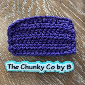 May include: A rectangular, hand-crocheted item in a vibrant purple hue. The chunky texture is visible, with a small white thread extending from the edge. The text "The Chunky Co by B" is displayed on a light blue and white label.