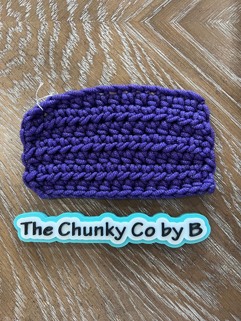 May include: A rectangular, hand-crocheted item in a vibrant purple hue. The chunky texture is visible, with a small white thread extending from the edge. The text "The Chunky Co by B" is displayed on a light blue and white label.