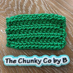 May include: A vibrant green, chunky crocheted rectangle with a dense, textured surface. The stitches are clearly defined. Beneath it is a label in white and light blue, bearing the text "The Chunky Co by B".