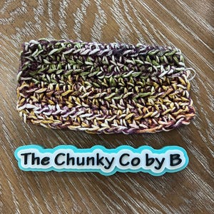 May include: A rectangular, hand-crocheted item in shades of purple, green, yellow, and white. The item is placed on a wooden surface. A blue and white label with the text "The Chunky Co by B" is below the crocheted item.