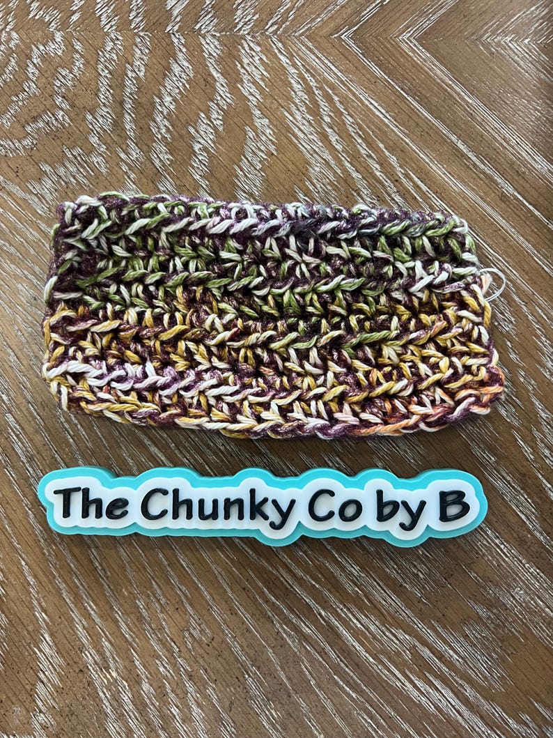May include: A rectangular, hand-crocheted item in shades of purple, green, yellow, and white. The item is placed on a wooden surface. A blue and white label with the text "The Chunky Co by B" is below the crocheted item.