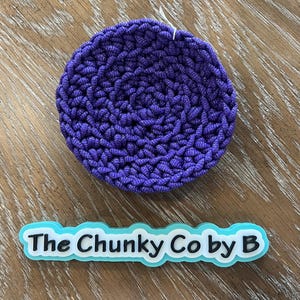 May include: A circular, deep purple crocheted item with a textured surface. Below it is a light blue and white sign that reads "The Chunky Co by B". The item is likely a coaster or trivet.
