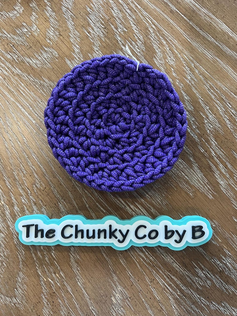 May include: A circular, deep purple crocheted item with a textured surface. Below it is a light blue and white sign that reads "The Chunky Co by B". The item is likely a coaster or trivet.