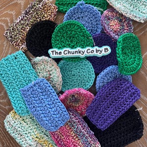 May include: A collection of handmade crochet items in various shapes and colors, including green, blue, purple, and multi-colored pieces. The items are arranged on a wooden surface, with the text "The Chunky Co by B" visible.