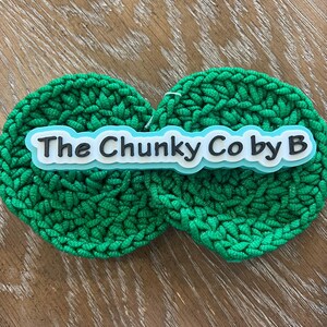 May include: Two round, bright green crocheted items with the text "The Chunky Co by B" in white letters with a black outline, set against a light wood grain surface. The crochet has a textured, chunky appearance.