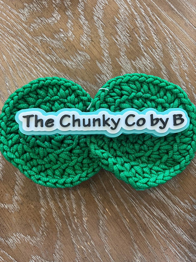 May include: Two round, bright green crocheted items with the text "The Chunky Co by B" in white letters with a black outline, set against a light wood grain surface. The crochet has a textured, chunky appearance.