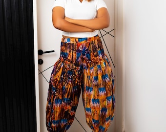 Puede incluir: Una blusa blanca de manga corta con cuello en V combinada con pantalones voluminosos de talle alto que presentan un estampado abstracto multicolor en tonos naranja, azul y marrón. Los pantalones tienen una silueta abullonada y sandalias doradas.