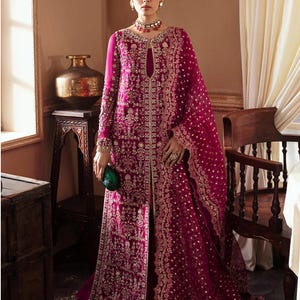 Puede incluir: Un vestido formal magenta de manga larga con intrincados bordados y adornos dorados. El vestido presenta un escote de ojo de cerradura y se combina con un dupatta a juego. Se sostiene un pequeño clutch verde en la mano.