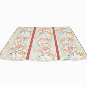May include: A folded, floral-patterned textile with a light green background and pink and orange flower designs. The textile has three panels with a vertical red stripe down the center of each panel. The edges are also trimmed in red.