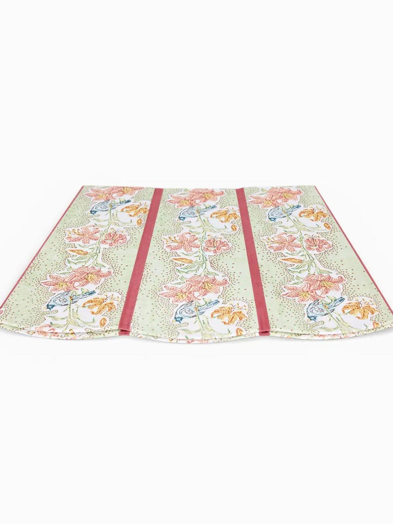 May include: A folded, floral-patterned textile with a light green background and pink and orange flower designs. The textile has three panels with a vertical red stripe down the center of each panel. The edges are also trimmed in red.