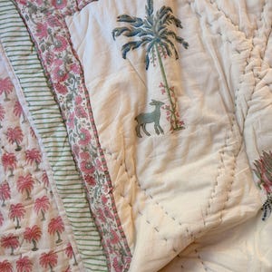May include: A white quilted blanket with a palm tree and deer design. The blanket features pink and green floral and striped borders. The palm tree is blue and green, and the deer is gray. The blanket is made of soft fabric.