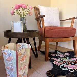 May include: A decorative wastebasket with a floral and bird pattern in shades of green, orange, and cream. The wastebasket is next to a wooden side table with a vase of pink flowers and a framed picture. A wooden chair with a red cushion and a patterned rug are also in the room.