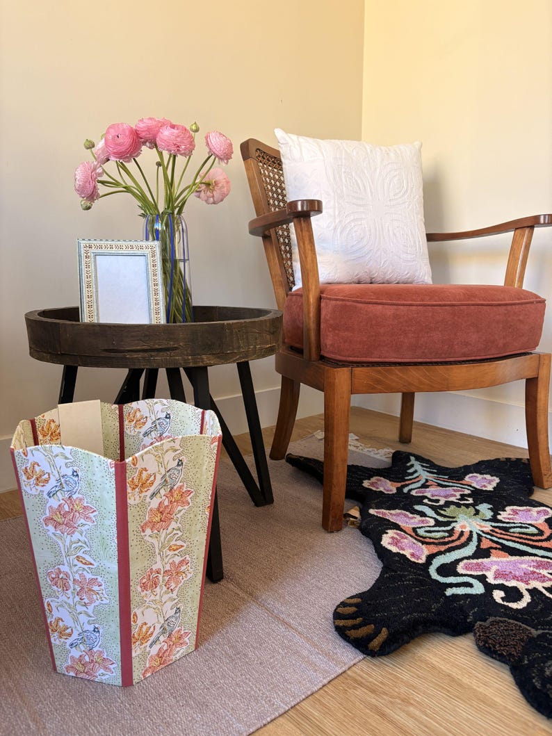 May include: A decorative wastebasket with a floral and bird pattern in shades of green, orange, and cream. The wastebasket is next to a wooden side table with a vase of pink flowers and a framed picture. A wooden chair with a red cushion and a patterned rug are also in the room.