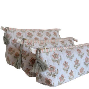 May include: Three white floral print cosmetic bags with gray tassels. The bags feature a repeating pattern of pink flowers and green leaves. Each bag has a zipper closure and a striped accent.