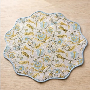 Puede incluir: Un mantel individual redondo con borde festoneado acabado en ribete azul claro. La tela presenta un fondo gris claro con un patrón repetitivo de flores y enredaderas en tonos amarillo mostaza y azul claro, creando un diseño botánico.