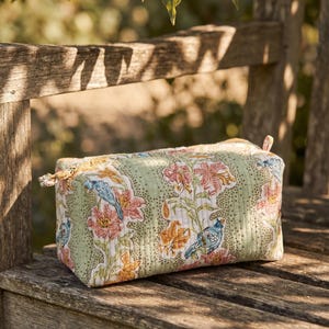 May include: A rectangular, quilted cosmetic bag with a floral and bird pattern in shades of green, pink, orange, and blue. The bag is sitting on a wooden bench in natural light. The bag has a zipper closure.