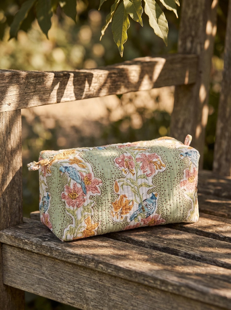 May include: A rectangular, quilted cosmetic bag with a floral and bird pattern in shades of green, pink, orange, and blue. The bag is sitting on a wooden bench in natural light. The bag has a zipper closure.