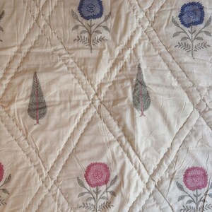 Può includere: Un tessuto trapuntato color crema caratterizzato da un motivo a rombi. Il tessuto è decorato con motivi stampati a blocchi di fiori blu e rosa, insieme a cipressi grigi. Un bordo decorativo presenta dettagli floreali di colore rosa.