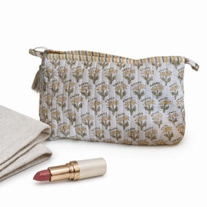 May include: A white quilted cosmetic bag with a yellow floral pattern, a beige tassel, and a striped top. A folded beige cloth and a lipstick with a pink hue are next to the bag.