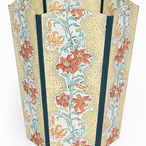 May include: A decorative wastebasket with a floral design. The wastebasket features a cream-colored background with a pattern of orange lilies and green leaves. Dark teal vertical stripes frame the floral pattern.