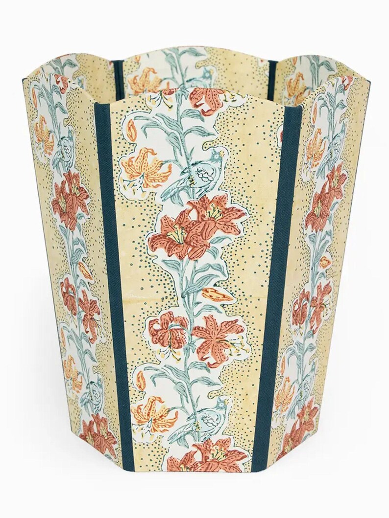 May include: A decorative wastebasket with a floral design. The wastebasket features a cream-colored background with a pattern of orange lilies and green leaves. Dark teal vertical stripes frame the floral pattern.
