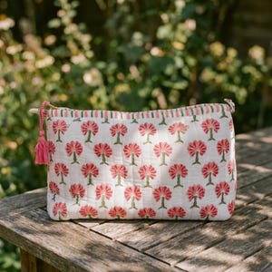 May include: A white cosmetic bag with a repeating pattern of pink floral designs. The bag has a pink tassel and a striped border. The bag is sitting on a wooden table in front of a blurred green background.