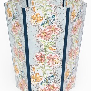 May include: A tall, decorative wastebasket with a floral and bird pattern. The wastebasket features a light blue background with a repeating pattern of pink and orange lilies, green leaves, and blue birds. Dark blue vertical stripes accent the sides.