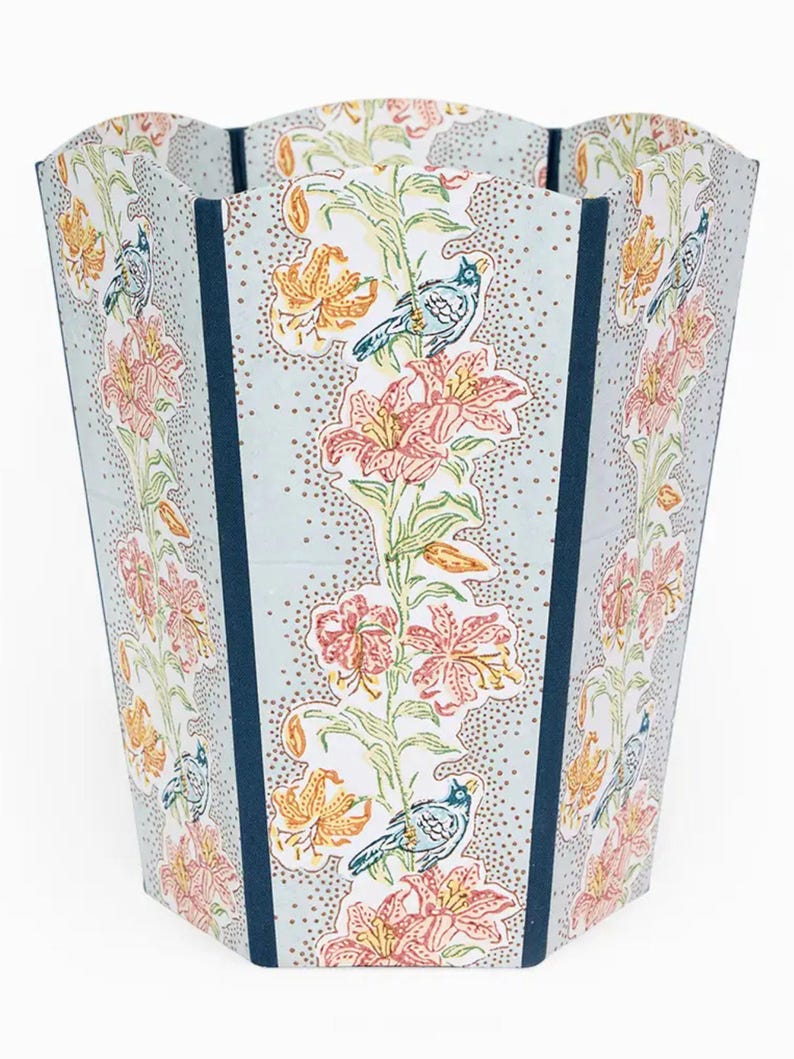 May include: A tall, decorative wastebasket with a floral and bird pattern. The wastebasket features a light blue background with a repeating pattern of pink and orange lilies, green leaves, and blue birds. Dark blue vertical stripes accent the sides.