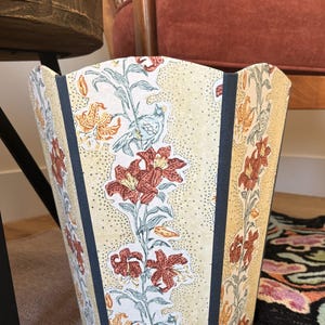 May include: A decorative wastebasket with a floral pattern. The wastebasket features a cream-colored background with a repeating pattern of red lilies, orange accents, and blue birds. Dark blue vertical stripes frame the floral design.
