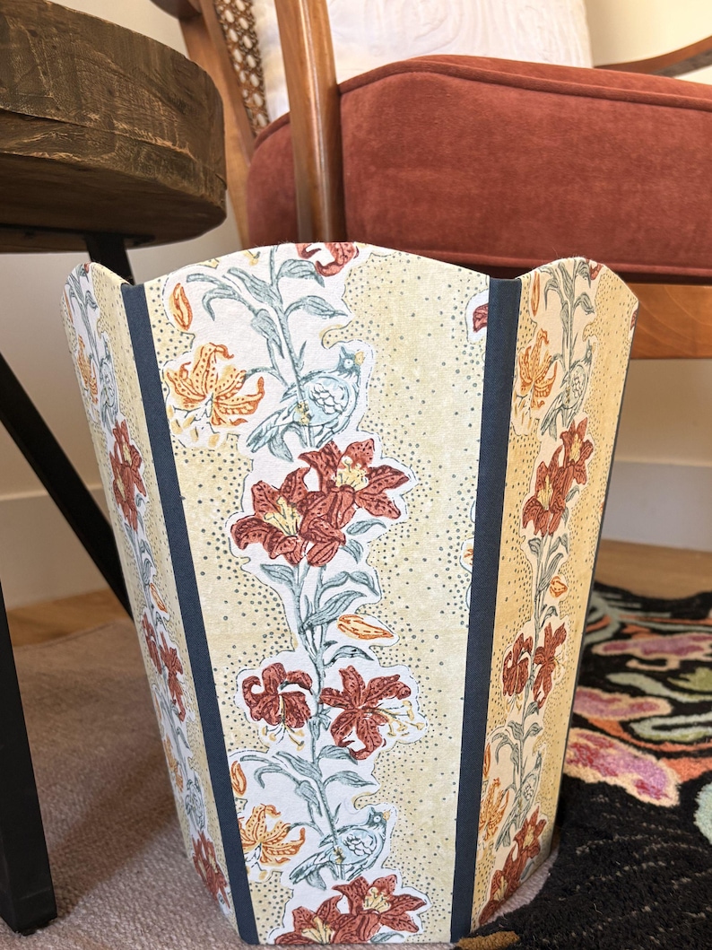 May include: A decorative wastebasket with a floral pattern. The wastebasket features a cream-colored background with a repeating pattern of red lilies, orange accents, and blue birds. Dark blue vertical stripes frame the floral design.