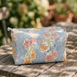May include: A rectangular, light blue cosmetic bag with a floral pattern of orange and pink lilies and blue birds. The bag is quilted and has a zipper closure. The bag is sitting on a wooden table.