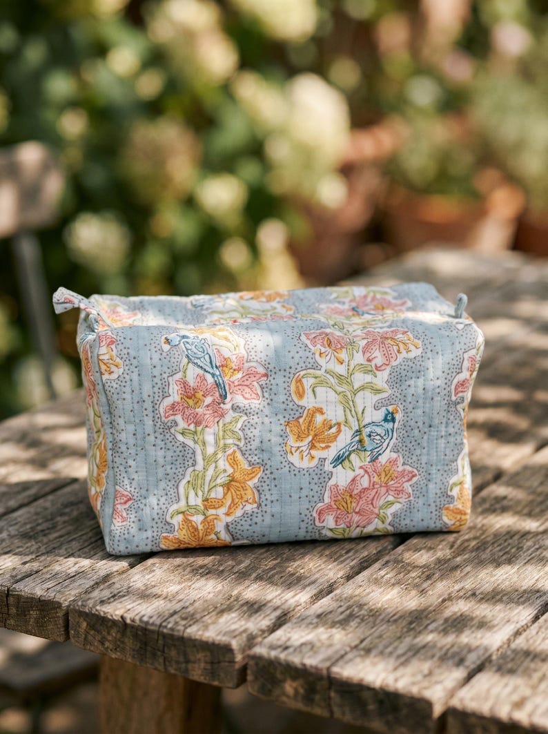 May include: A rectangular, light blue cosmetic bag with a floral pattern of orange and pink lilies and blue birds. The bag is quilted and has a zipper closure. The bag is sitting on a wooden table.