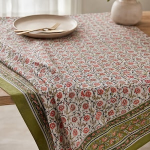 Puede incluir: Un mantel con estampado floral con fondo crema y diseños de flores rojas. El mantel tiene un borde verde y está colocado sobre una mesa de madera. Un plato y cubiertos están sobre la mesa.