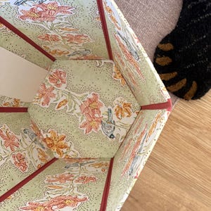 May include: A hexagonal storage box with a floral and bird pattern in shades of orange, green, and white. The edges are trimmed with a dark red binding. The interior and exterior feature the same design.